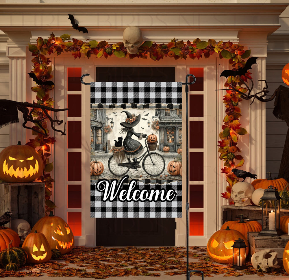 A perfect Halloween Garden Flag featuring an adorable Witch on her bicycle with her black cat, riding down a street decorated for Halloween.