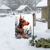 A Red and Rust Colored Doberman Pinscher Dog in a Winter Wonderland setting, wearing a festive collar of greenery and berries with pine cones.