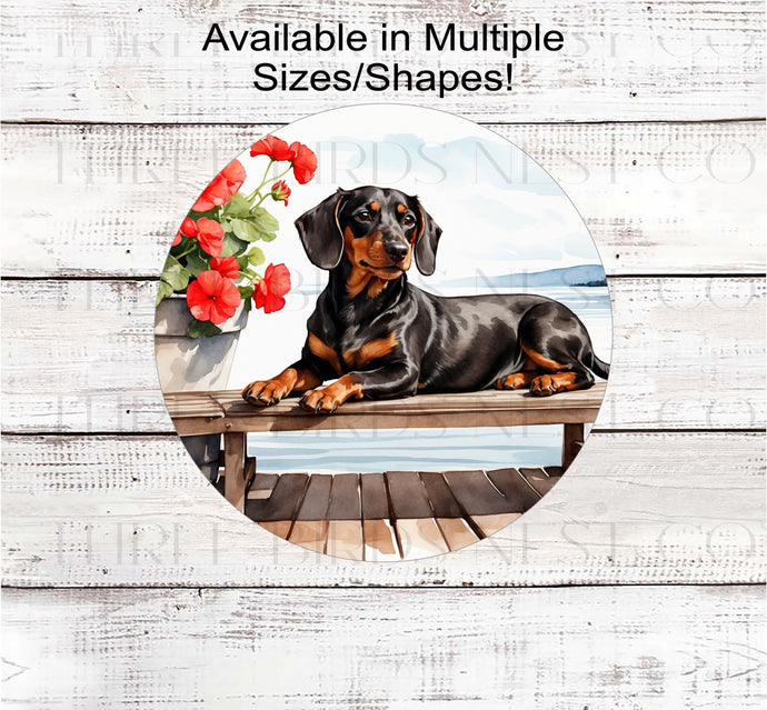 A black Dachshund Dog on a dock with a flower pot overlooking a lake.