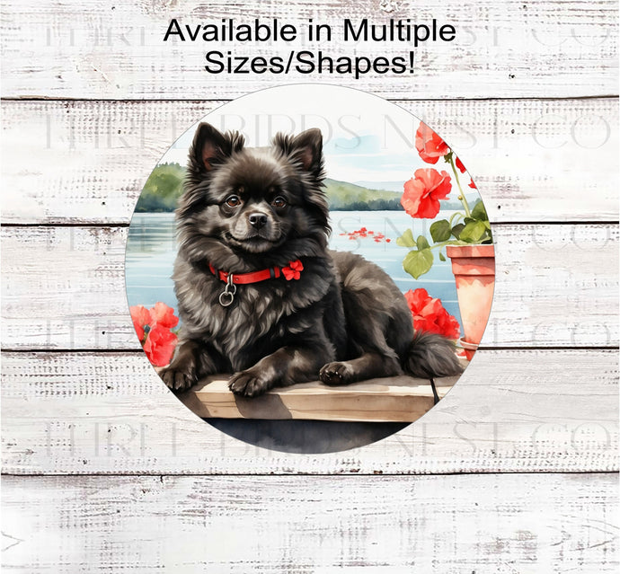 A black Pomeranian Dog on a dock with a flower pot overlooking a lake.