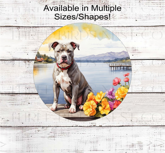 A grey and white Pit Bull Dog on a dock with a flower pot overlooking a lake.